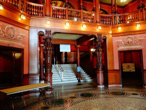 Interior of a grand building featuring ornate wooden columns, a staircase, and decorative tile flooring.