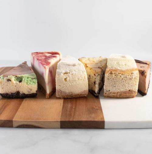 A variety of cheesecake slices displayed on a wooden cutting board, featuring different flavors and colors.