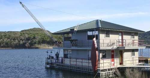 A two-story houseboat on a lake, featuring a green roof and a crane for lifting items. Surrounding hills in the background.