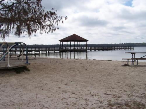A sandy beach with a wooden pier and gazebo extending over calm water, under a cloudy sky.