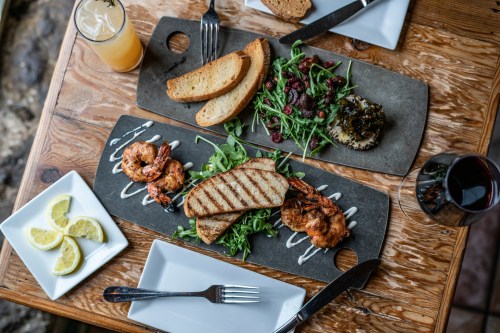 Two plates of food on a wooden table: grilled shrimp with bread, salad, and lemon slices, alongside a drink.
