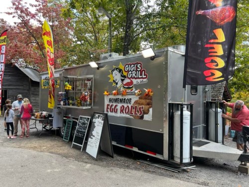 A food truck named "Gigi's Grill" serving homemade egg rolls, surrounded by trees and people enjoying the outdoor setting.