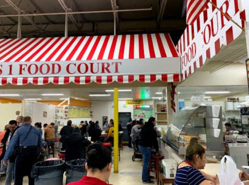 A bustling food court with red and white striped awning, featuring diners at tables and food vendors in the background.
