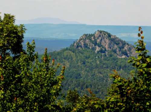 A rocky peak rises above a lush green forest, with distant mountains visible under a clear sky.