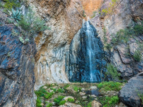 A serene waterfall cascading down rocky cliffs, surrounded by lush greenery and rugged terrain.