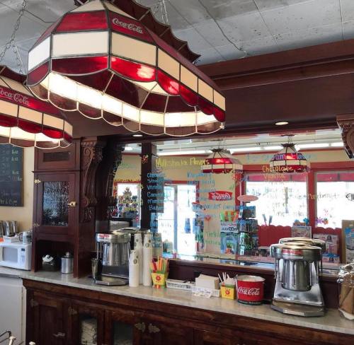 A vintage ice cream shop counter with red and white lamps, a menu on the mirror, and ice cream machines.