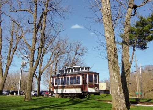 A vintage tram on a grassy area surrounded by bare trees under a clear blue sky.