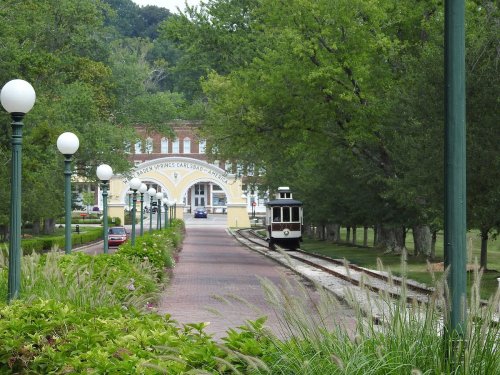 A scenic pathway lined with trees and lamps, leading to a historic archway and a vintage streetcar on the tracks.