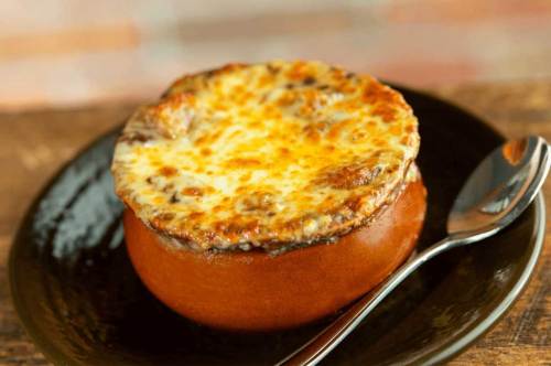 A bowl of French onion soup topped with melted cheese, served on a dark plate with a spoon beside it.