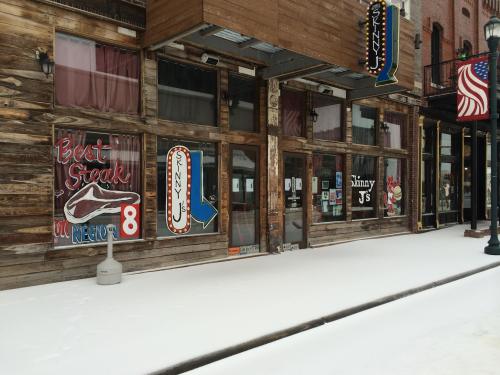 A rustic restaurant exterior with wooden walls, colorful signs, and a snowy sidewalk in front.