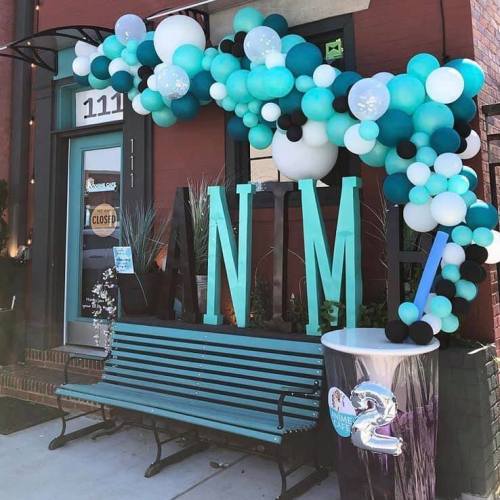 A colorful balloon arch in teal, black, and white decorates a storefront entrance with a bench and large letters "NM."