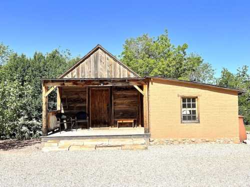 A rustic wooden cabin with a porch, surrounded by greenery and gravel, under a clear blue sky.