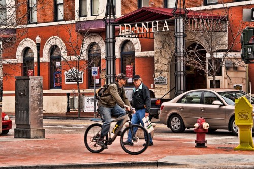 A cyclist rides past a historic building with pedestrians and cars on a city street.