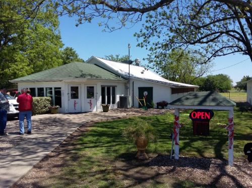 A quaint building with a green roof, open sign, and people outside in a grassy area on a sunny day.