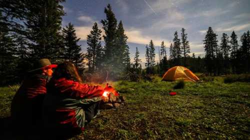 A couple sits by a campfire under a starry sky, with a tent and trees in the background.