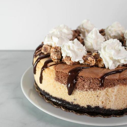 A delicious cheesecake topped with whipped cream, chocolate drizzle, and cookie pieces on a white plate.