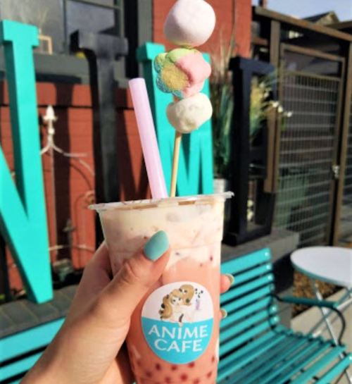 A hand holding a colorful drink topped with marshmallows, in front of a vibrant "ANIME CAFE" sign.