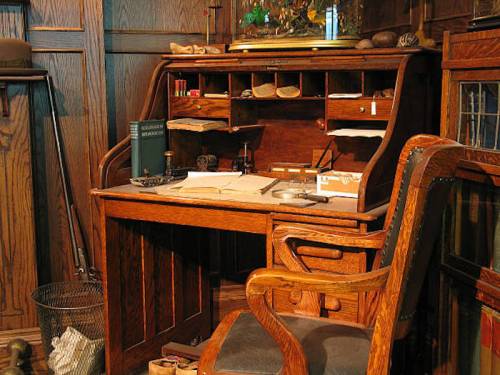 A vintage wooden desk with an ornate chair, papers, and various items arranged on and around it in a cozy room.