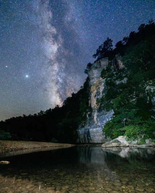 A serene night scene featuring the Milky Way over a rocky cliff and calm water, surrounded by lush trees.