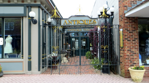 A decorative gate with "Antiques" sign leads into a quaint shop, surrounded by brick pathways and storefronts.