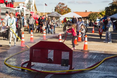 A community event with people using hoses, children playing, and tents in the background, set on a sunny day.