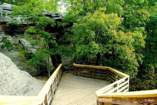 A wooden walkway curves through lush greenery and rocky terrain, leading to a natural stone formation.