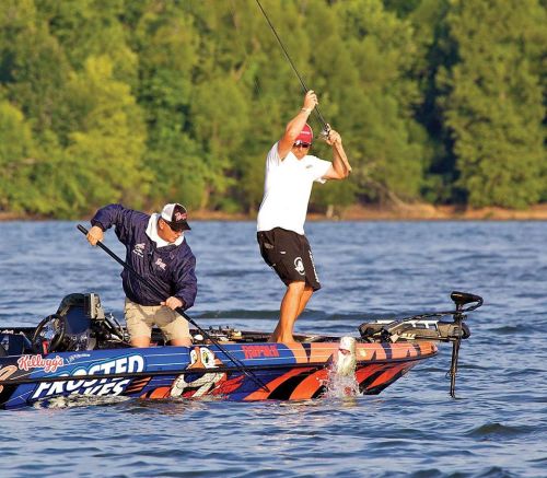 Two men on a fishing boat, one casting a line while the other assists, with trees and water in the background.