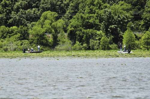 Two boats on a lake surrounded by lush green trees and lily pads, with people fishing or enjoying the outdoors.