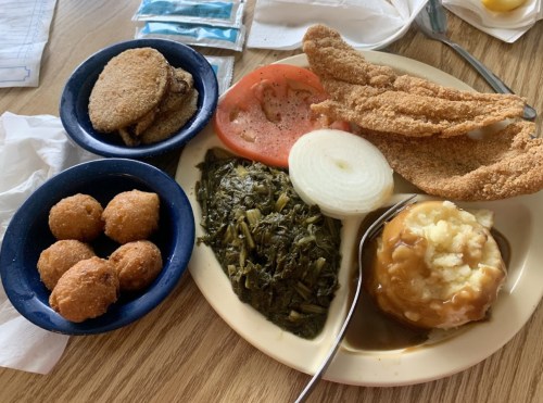 A plate of fried fish, collard greens, mashed potatoes with gravy, sliced onion, tomato, and hushpuppies.