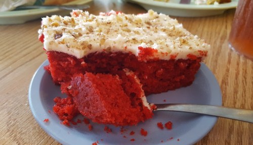 A slice of red velvet cake with cream cheese frosting and chopped nuts on a blue plate, with a fork beside it.