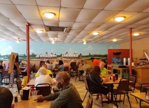 A busy diner with patrons seated at tables, featuring a mural of a rural landscape on the wall.