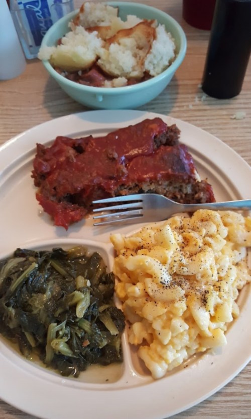 A plate with meatloaf topped with sauce, macaroni and cheese, and greens, with a bowl of mashed potatoes in the background.