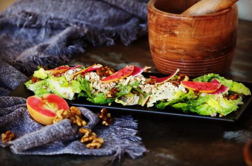 A vibrant salad with lettuce, nuts, and pink radish, served on a black plate with a wooden bowl and apple nearby.
