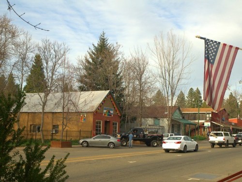 A small town street scene with cars, colorful buildings, and an American flag waving in the foreground.