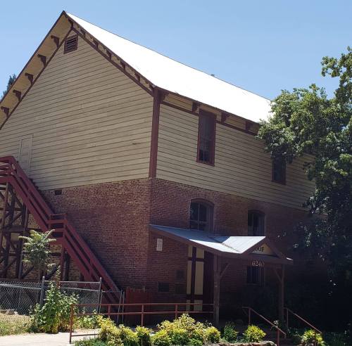 Historic building with a wooden upper structure and brick lower level, surrounded by greenery and a clear blue sky.