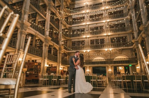 A couple embraces in a grand library with tall shelves, elegant decor, and soft lighting, creating a romantic atmosphere.