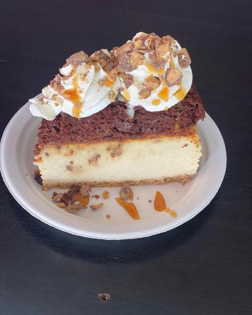 A slice of layered cake with cheesecake, chocolate cake, whipped cream, and caramel drizzle, served on a white plate.