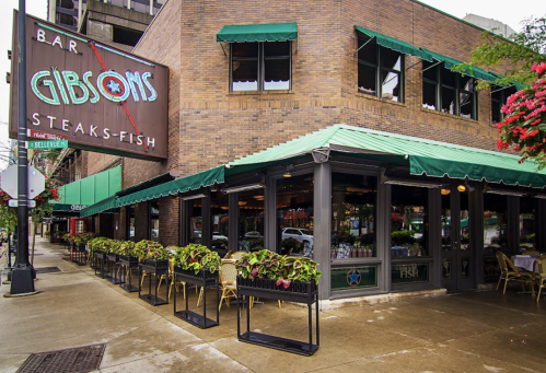 Exterior of Gibsons Bar, featuring a green awning, outdoor seating, and a sign for steaks and fish.