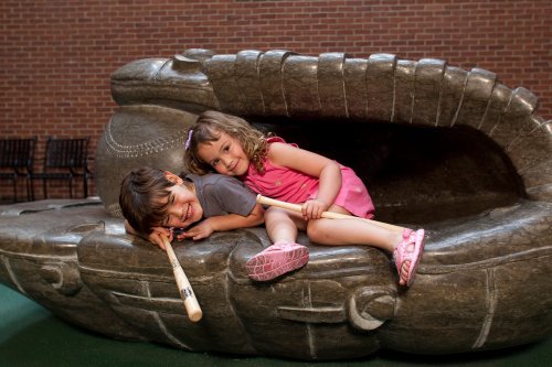 Two children play on a large stone sculpture, one sitting inside while the other leans on it, both holding sticks.