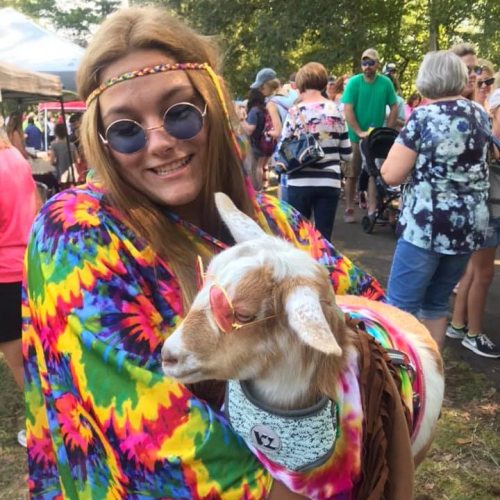 A person in colorful tie-dye attire holds a goat wearing sunglasses, surrounded by a lively outdoor crowd.
