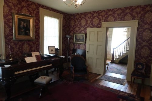 A vintage room with a piano, ornate wallpaper, and a staircase visible through an open door.