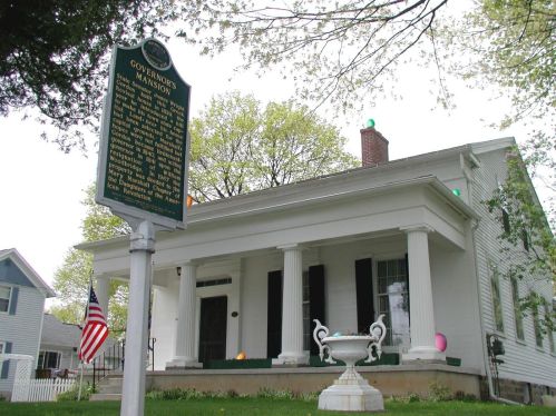 Historic Governor's Mansion with a green sign, white columns, and an American flag in front. Trees in the background.