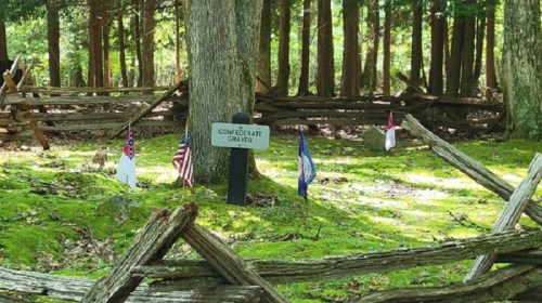 A sign reading "Confederate Graves" near a wooded area, surrounded by flags and a rustic wooden fence.