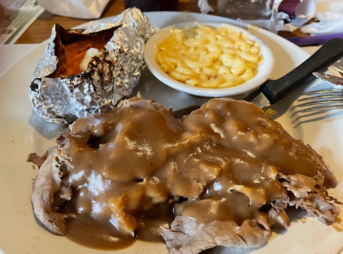 A plate with sliced roast beef covered in gravy, a side of macaroni and cheese, and a baked potato wrapped in foil.