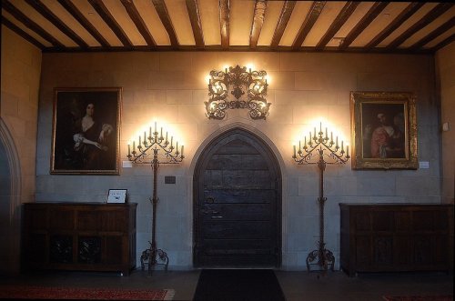 A dimly lit hallway featuring two ornate candelabras, paintings, and a large wooden door with an arched top.