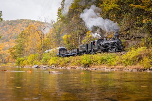 A vintage steam train travels along a river, surrounded by colorful autumn foliage and rolling hills.