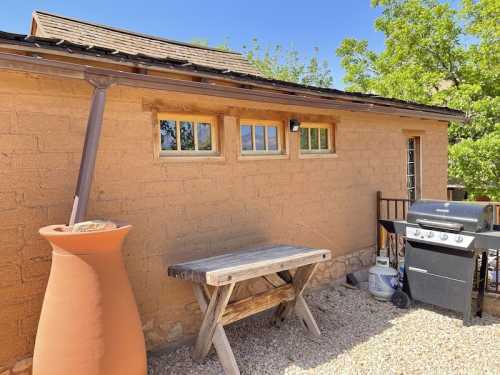 A rustic outdoor space featuring a clay pot, wooden table, and a grill against a textured wall and clear blue sky.