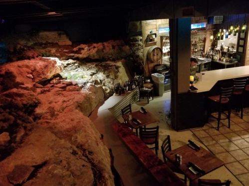 A dimly lit restaurant featuring a rocky wall, bar area, and wooden tables with chairs.