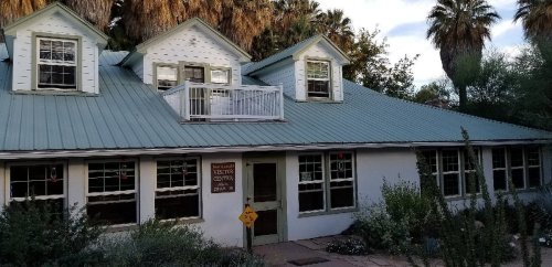A charming white building with a green metal roof, surrounded by palm trees and desert plants, featuring large windows.