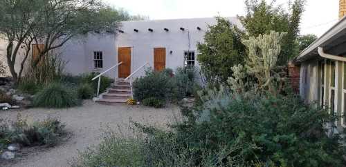 A desert-style home with wooden doors, surrounded by lush greenery and cacti in a gravel landscape.